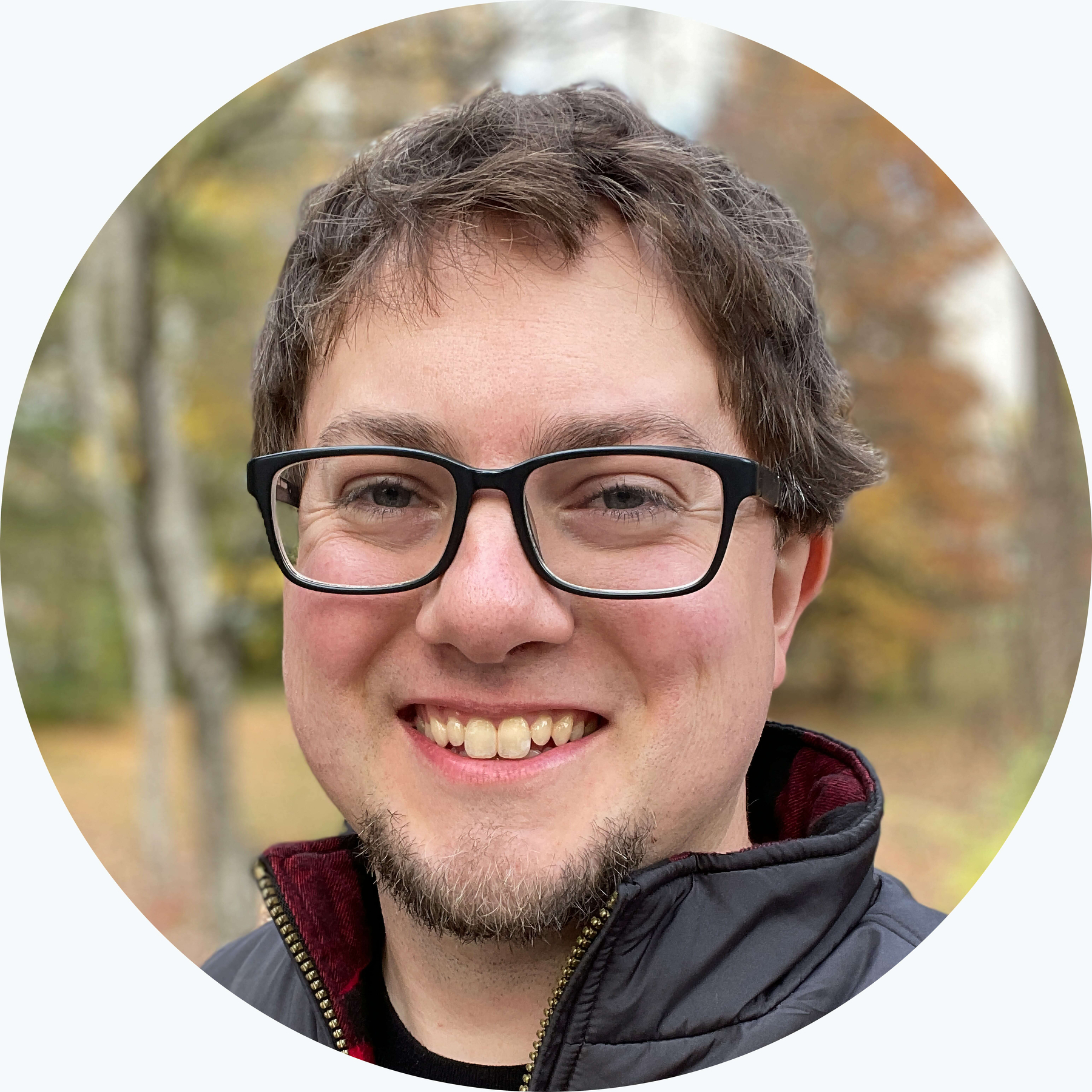 Smiling man with medium length brown hair, glasses, and a trimmed beard, wearing a black jacket outdoors with autumn foliage in the background.